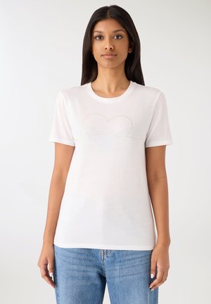 Giovane donna con lunghi capelli scuri che indossa una maglietta bianca con un cuore dorato tenue e la parola "love" abbinata a un paio di jeans azzurri.