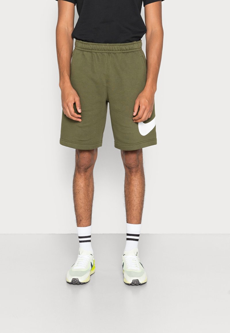 Olivgrüne Sportshorts mit einem elastischen Bund, seitlichen Taschen und einem auffälligen weißen Logo am linken Bein. Getragen mit weißen Socken und Turnschuhen.
