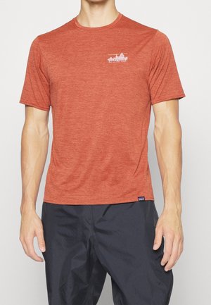 Mann trägt ein rötlich-oranges kurzärmeliges Patagonia T-Shirt mit kleinem weißem Logo und schwarze Hose, steht vor einem schlichten hellen Hintergrund.