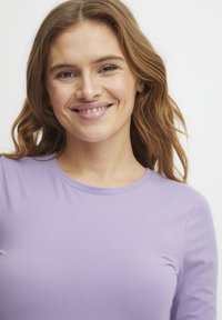 Licht paarse, lange mouwen shirt van zachte stof met een ronde halslijn. Model glimlacht met golvend, schouderlang haar.