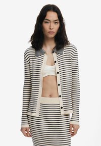 Cardigan a righe in crema con linee blu navy, dotato di colletto, chiusura a bottoni e polsini a coste, abbinato a una gonna coordinata.