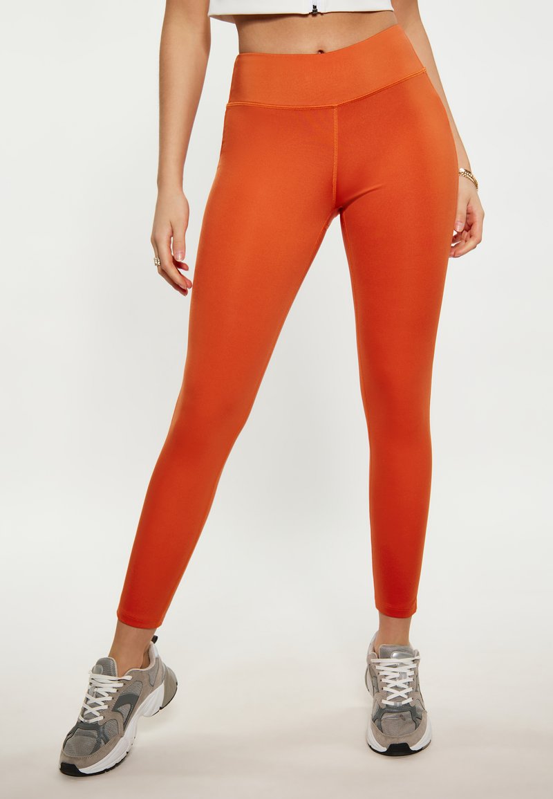 Oranje hoge tailleleggings met een gladde afwerking en elastische tailleband, gecombineerd met grijze sportschoenen. Volledige lengte en aansluitend ontwerp.