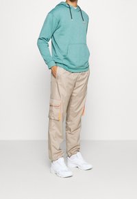 Felpa cappuccio turchese con tasca frontale, abbinata a pantaloni cargo beige con dettagli arancioni e tasche. Scarpe da ginnastica bianche.