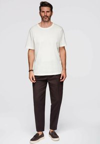 Muž stojící oblečený v jednoduché bílé košili s krátkým rukávem, tmavých elegantních kalhotách a tmavých slip-on botách s třásněmi na světlém pozadí.