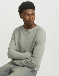 Hellgrauer gerippter Pullover mit langen Ärmeln und rundem Ausschnitt, kombiniert mit verwaschenen blauen Jeans. Model sitzt auf einem weißen Stuhl.