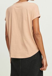 Mujer vestida con una camiseta beige de manga enrollada y una falda de mezclilla negra, mostrada desde la espalda con el brazo relajado a su lado.