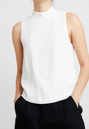 Kobieta ubrana w bezrękawnik z białym golfem, włożony do czarnych spodni z wysokim stanem, z rękami w kieszeniach.