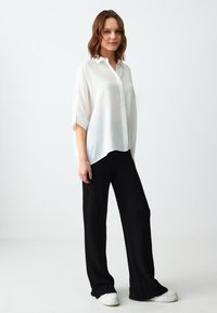 Chemise blanche à manches courtes avec col et détails aux poignets, associée à un pantalon noir texturé à larges jambes et des baskets blanches sur un fond uni.