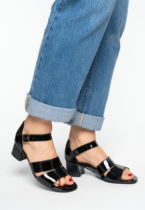 Fødder iført sorte block hælsandaler i laklæder med ankelstropper og rødt neglelak, kombineret med opkørte blå jeans.