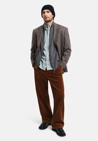 Man med en grå blazer över en grönrandig skjorta, bruna kordfloretbyxor och en svart mössa, med svarta skor. Avslappnad lagerlook.