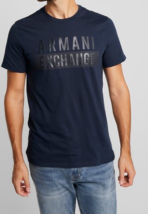 Mand iført navyblå Armani Exchange t-shirt og lyseblå jeans, stående foran en ensfarvet grå baggrund.