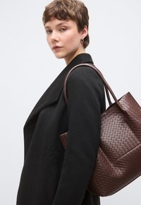 Καφέ υφαντή δερμάτινη τσάντα ώμου με δύο χειρολαβές, με δομημένο σχήμα και υφή επιφάνειας, συνδυασμένη με μαύρο παλτό.
