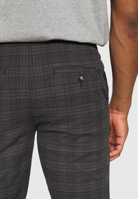 Pantaloni neri a quadri con tessuto strutturato, tasca posteriore con bottone e linee a contrasto marroni e grigie nel motivo.