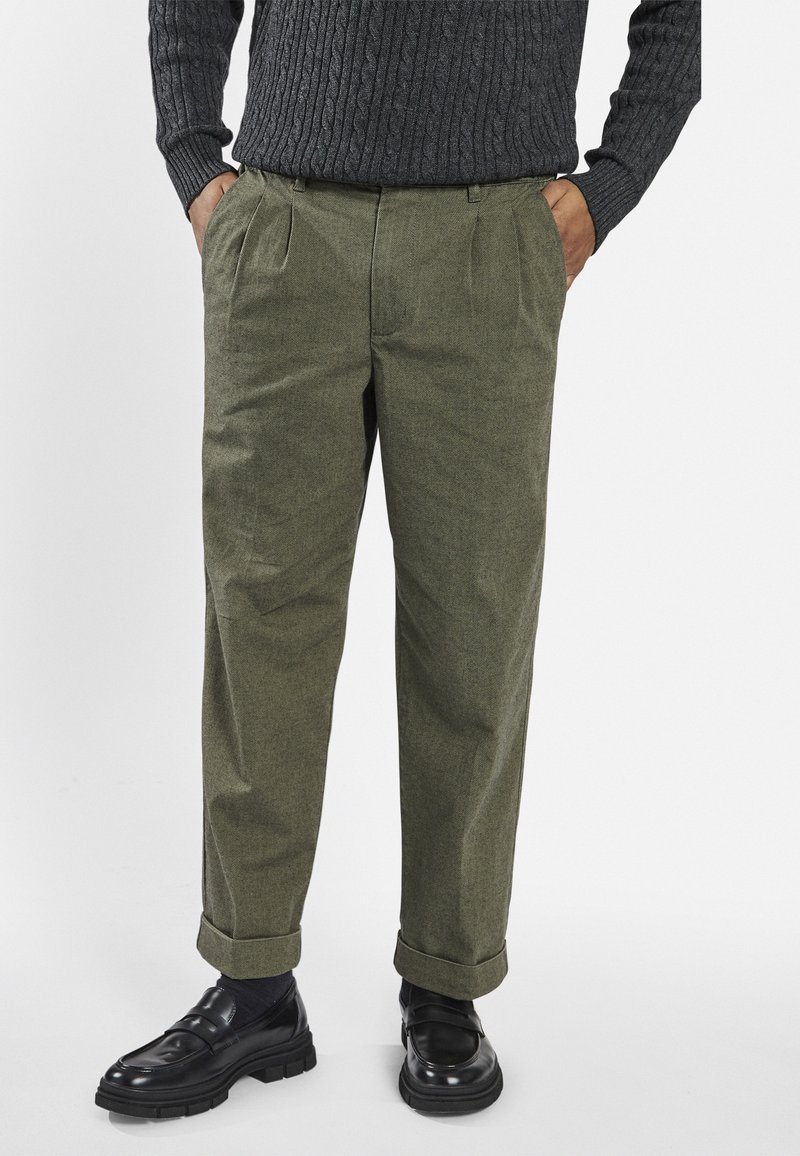 Pantalon vert olive en tissu texturé, avec pli creux à l'avant et revers remontés. Associé à des mocassins en cuir noir.