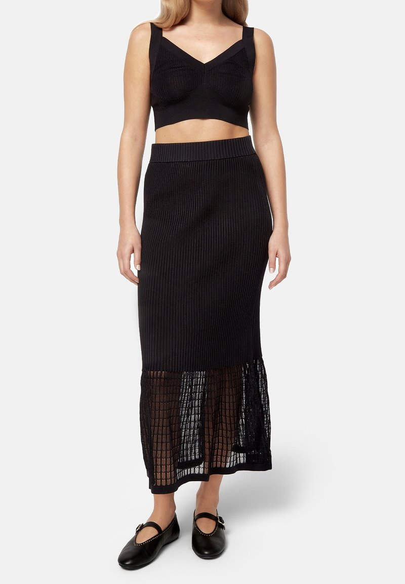 Femme portant un crop top côtelé noir et une longue jupe assortie avec un panneau transparent, associée à des ballerines noires style Mary Jane.