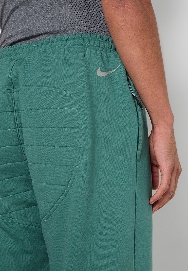 Persona che indossa pantaloni sportivi imbottiti verdi con un logo Nike grigio, una tasca con zip laterale e una parte superiore in rete grigia visibile in vita.