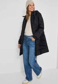 Sort puffersjakke med hætte og lynlås, beige sweater, blå brede jeans og hvide sneakers. Enkel design med quiltet tekstur.