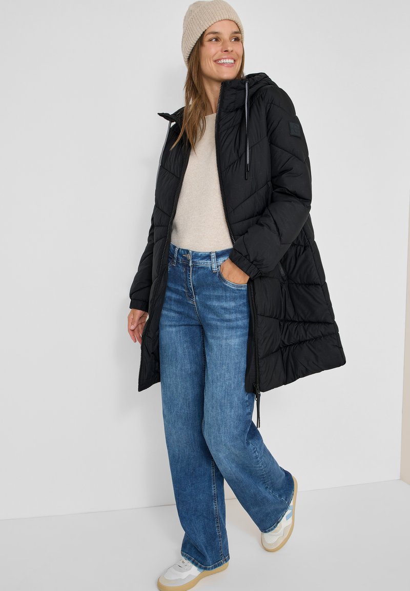 Sort puffersjakke med hætte og lynlås, beige sweater, blå brede jeans og hvide sneakers. Enkel design med quiltet tekstur.