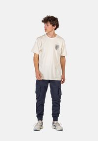 Beige Baumwoll-T-Shirt mit kurzen Ärmeln und Grafik auf der Brust; kombiniert mit marineblauen Cargo-Hosen mit Seitentaschen und eng zulaufenden Bündchen, getragen mit Sneakers.