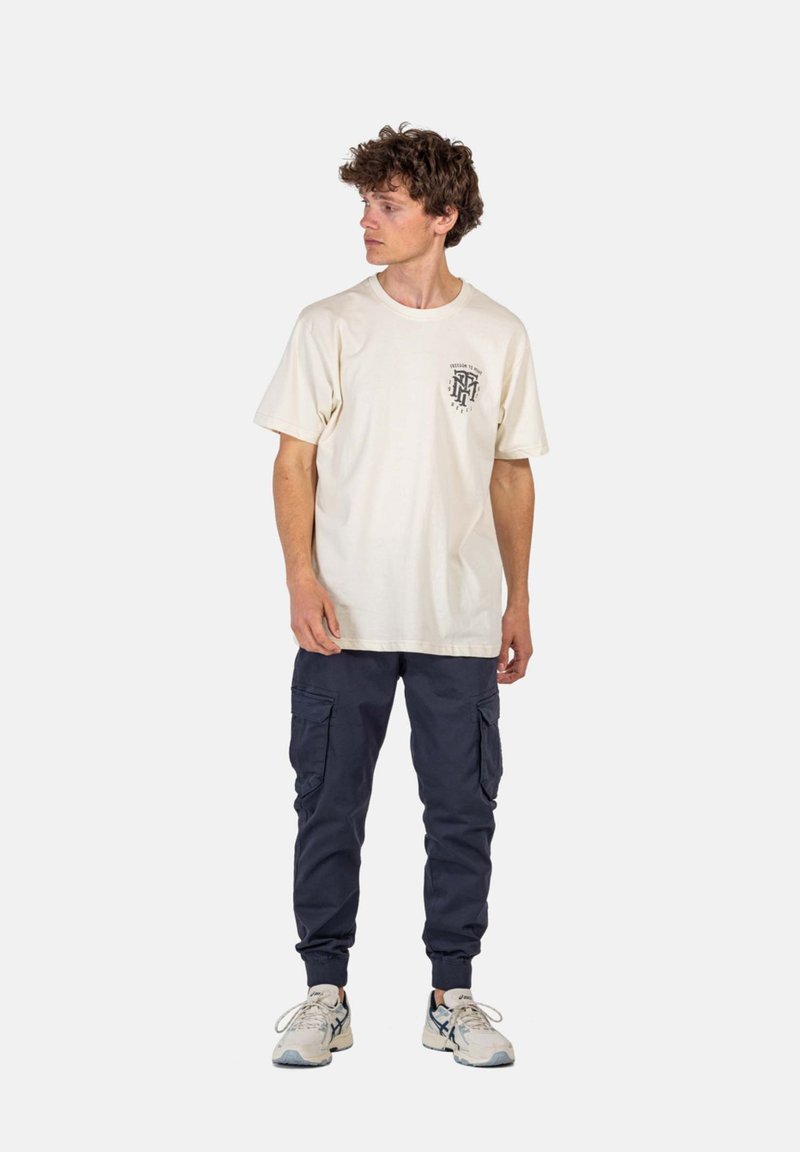 Beige Baumwoll-T-Shirt mit kurzen Ärmeln und Grafik auf der Brust; kombiniert mit marineblauen Cargo-Hosen mit Seitentaschen und eng zulaufenden Bündchen, getragen mit Sneakers.