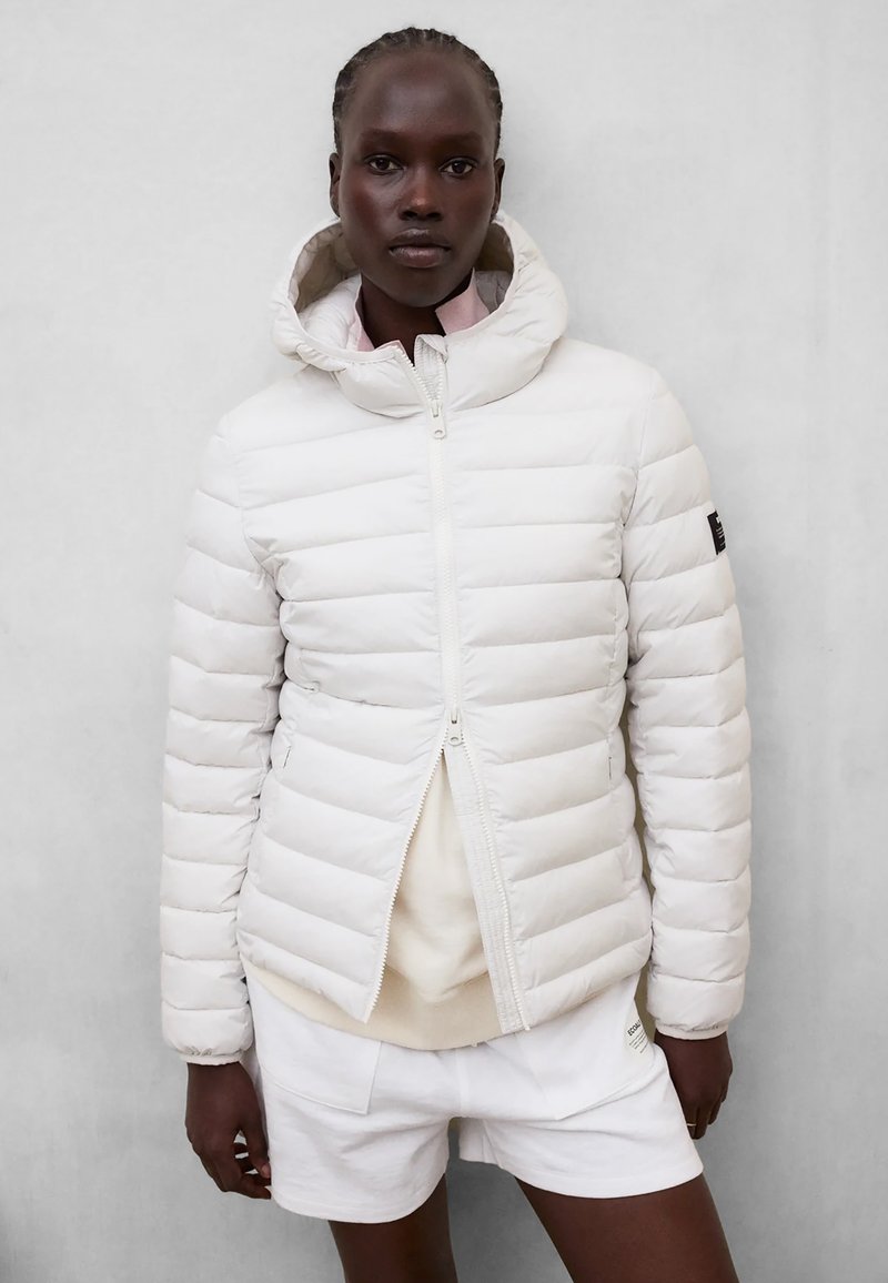Femme portant une veste matelassée blanche à capuche par-dessus un haut beige et un short blanc, debout devant un fond gris clair uni.