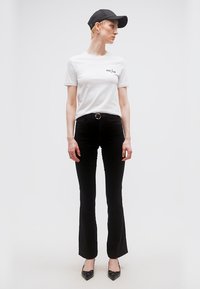 Γυναίκα που φοράει λευκό μπλουζάκι με γραφικά, μαύρο φαρδύ παντελόνι με ζώνη, μαύρο καπέλο και μαύρες μυτερές γόβες, στέκεται μπροστά σε λευκό φόντο.