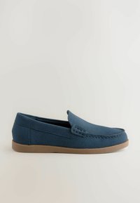 Mocassins en daim bleu avec un bout arrondi, une couture décorative et une semelle en caoutchouc brun, présentant un profil bas et un design à enfiler.