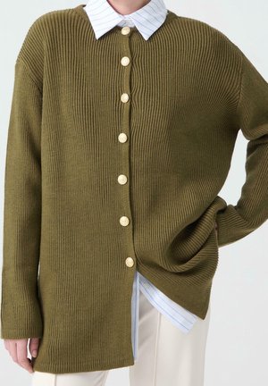 Personne portant un cardigan côtelé vert olive avec des boutons dorés sur une chemise blanche à rayures bleues et un pantalon clair.