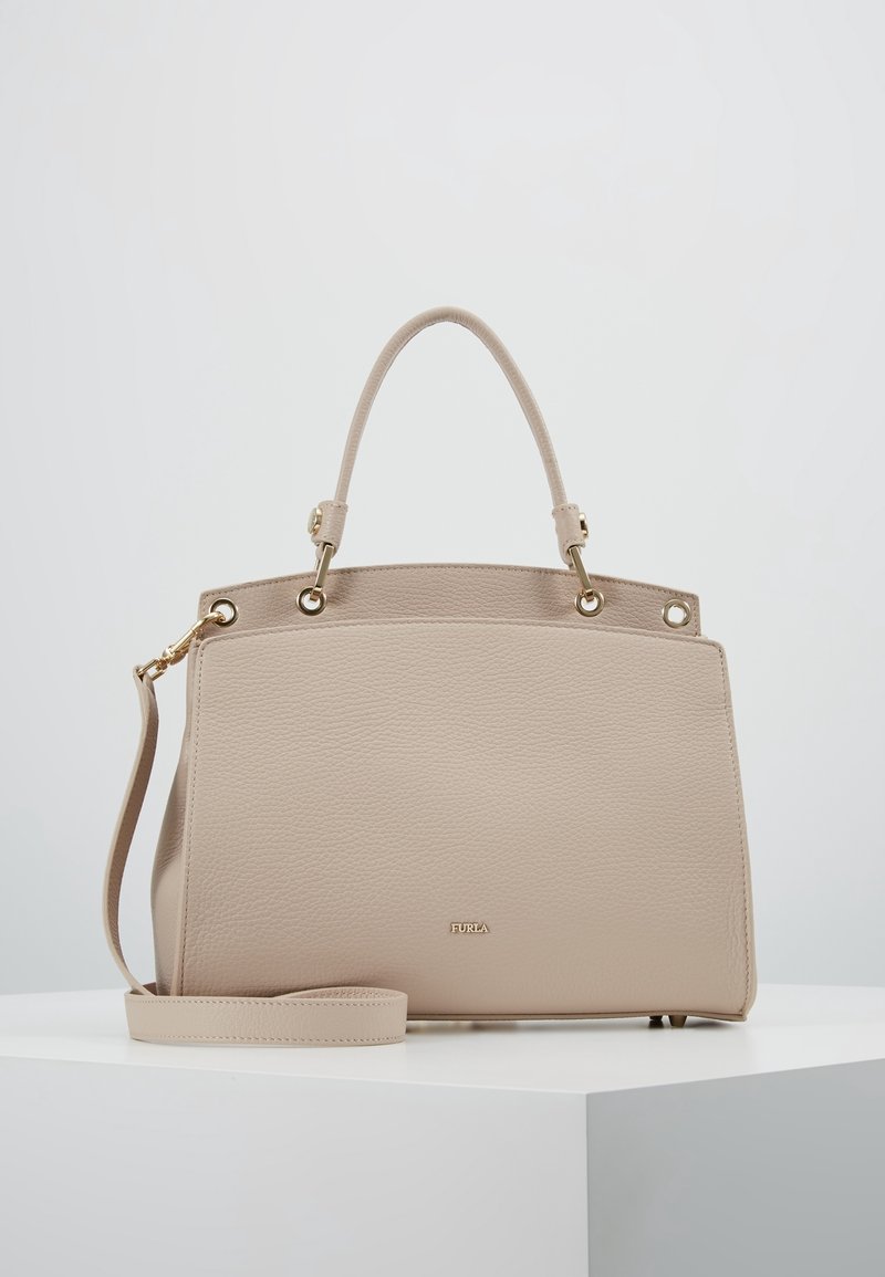 Sac à main en cuir beige avec poignées supérieures incurvées, bandoulière amovible, quincaillerie dorée et petit logo « Furla » sur le devant, posé sur une surface blanche.