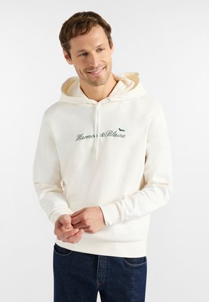 Lächelnder Mann, der einen weißen Hoodie mit dem Schriftzug "Harmon & Blaine" und einem kleinen Hundelogo trägt, kombiniert mit dunkelblauen Jeans, vor weißem Hintergrund.