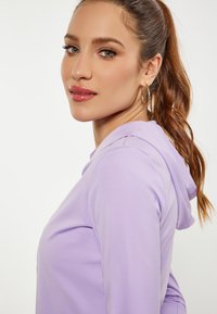 Felpa lilla con cappuccio e texture morbida. Presenta una vestibilità rilassata e una vista di profilo laterale di una persona con lunghi capelli mossi e orecchini a cerchio.