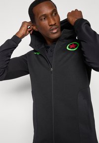 Čierna mikina na zips s vysokým golierom, s logom zelenej hviezdy, textúrovanými bočnými panelmi a hladkým povrchom látky.