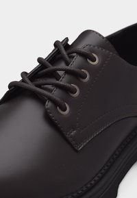 Μαύρο δερμάτινο παπούτσι φόρεμα με στρογγυλή μυτερή μύτη, διακοσμητικές ραφές και μαύρα κορδόνια που περνούν μέσα από τέσσερα μεταλλικά ρουχαλάκια σε κάθε πλευρά.