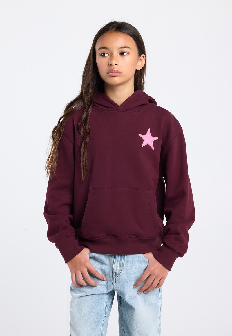 Adolescente debout, les mains près des poches, portant un sweat à capuche bordeaux avec une étoile rose sur la poitrine et un jean bleu clair, regardant sur le côté.