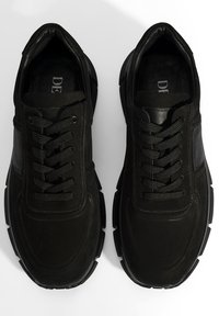 Schwarze Sneakers mit strukturierter Oberfläche, versehen mit glatten Lederakzenten, Gummisohlen und traditionellem Schnürdesign. Keine sichtbare Markenkennung.
