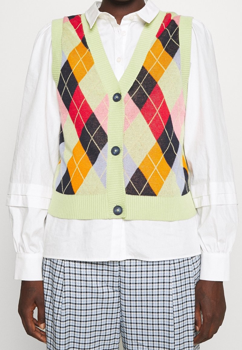 Gilet sans manches en tricot vert à losanges argyle orange, rouge, noir et rose, porté sur une chemise blanche boutonnée et un pantalon à carreaux.