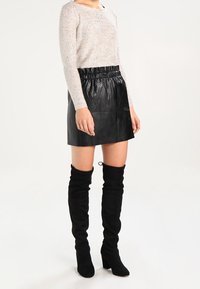 Top de manga larga beige claro con brillo, combinado con una minifalda de cuero negra y botas altas de ante negras con cintura fruncida.