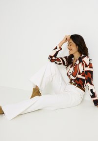 Camisa de botones con un atrevido patrón geométrico en rojo, negro y blanco; combinada con pantalones acampanados blancos y botas de tobillo en tono marrón. Materiales suaves y texturizados.