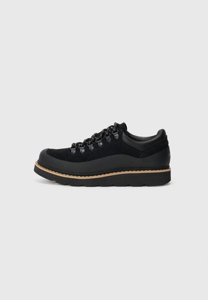 Black hiking-style sneaker with thick sole, metal eyelets, and contrast tan stitching on a plain white background.