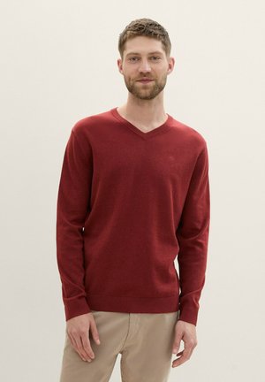 Maglione rosso a V in morbido tessuto lavorato a maglia. Presenta maniche lunghe e polsini a coste. Indossato con pantaloni chiari, sfondo semplice.