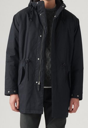 Mand iført sort hætteparka med lynlås og trykknapper over en mørkegrå sweater og bukser.