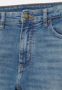 Gros plan sur un jean en denim bleu montrant la ceinture, le bouton avec le texte de la marque, les passants de ceinture et les coutures de la poche avant.