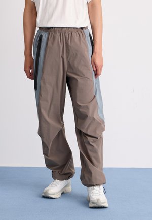 Personne portant un pantalon ample marron avec des panneaux latéraux gris et des poches zippées, associé à des baskets blanches sur un sol bleu.
