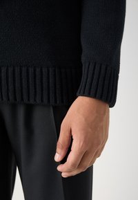 Main droite d'une personne portant un pull en maille noir et un pantalon noir, reposant à côté de la hanche sur un fond clair.