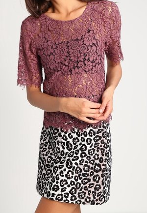 Kvinde iført en lilla blonde-top med korte ærmer og et sort-hvidt leopardprint nederdel, stående med hænderne foldet.