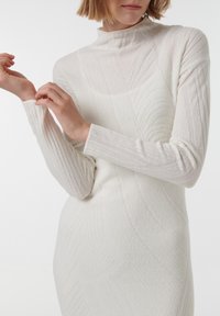 Gebreid jurkje in off-white met een hoge hals, geribbelde textuur en subtiele golfpatronen. Lange mouwen en een aansluitende silhoët.