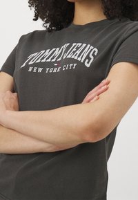 Camiseta de algodón gris oscuro con la inscripción "TOMMY JEANS NEW YORK CITY" en letras blancas, con un pequeño acento en rojo y azul. Manga corta.