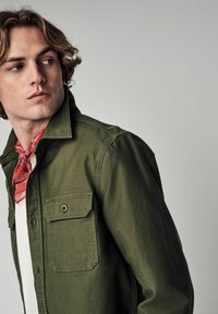 Olive green shirt jacket with a button closure and two chest pockets, paired with a red bandana around the neck and white shirt underneath.