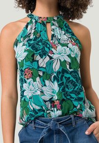 Haut sans manches à fleurs en bleu sarcelle, vert et blanc avec un col en forme d’œil-de-bœuf. Présente de grandes fleurs et feuilles sur un tissu léger.