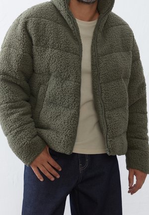 Man wearing olive green textured puffer jacket over beige shirt and dark blue jeans, standing with one hand resting on hip.